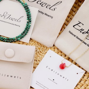 Op de afbeelding: Twee kleine witte zakjes met de tekst "Endi Jewels" erop gedrukt. E&eacute;n zakje bevat een groene malachiet kralenketting. Het andere zakje bevat een gouden ketting met een rode edelsteen hanger. De zakjes liggen op een geweven rieten oppervlak.