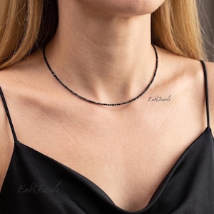May include: A delicate black beaded necklace. The necklace is worn around the neck and is composed of small, evenly spaced beads. The model is wearing a black spaghetti strap top. The text "Endi Jewels" is visible on the chest.