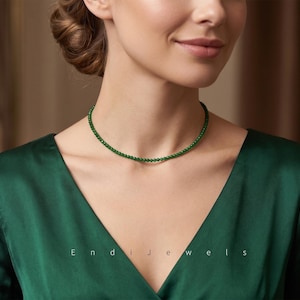 May include: A delicate choker necklace featuring small, uniform, round green beads. The necklace rests against the neck, paired with a deep V-neck emerald green satin garment. The text Endi Jewels is visible in white lettering below the necklace.