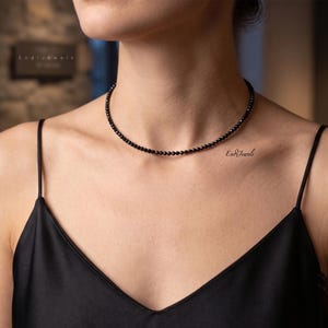 K&ouml;nnte beinhalten: Eine zierliche schwarze Perlenhalskette als Choker, bestehend aus kleinen, gleichm&auml;&szlig;igen, facettierten runden Perlen, die auf einem einzelnen Strang aufgereiht sind. Die Kette liegt eng am Hals an und jede Perle hat eine dunkle Oberfl&auml;che.