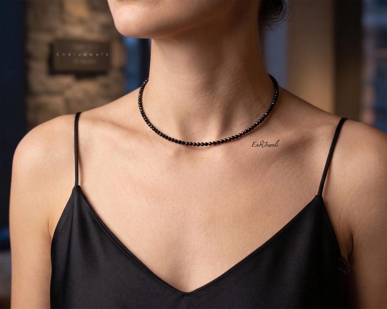 K&ouml;nnte beinhalten: Eine zierliche schwarze Perlenhalskette als Choker, bestehend aus kleinen, gleichm&auml;&szlig;igen, facettierten runden Perlen, die auf einem einzelnen Strang aufgereiht sind. Die Kette liegt eng am Hals an und jede Perle hat eine dunkle Oberfl&auml;che.