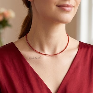 May include: A delicate red beaded necklace. The necklace is worn on a person with a red v-neck top. The necklace is a choker style, with small, round beads. The background is blurred, with a vase of flowers visible.