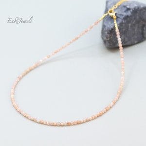 K&ouml;nnte beinhalten: Eine zarte goldene Kettenkette mit kleinen, hellrosa Sunstone-Perlen. Die Kette wird auf einem grauen Stein pr&auml;sentiert.