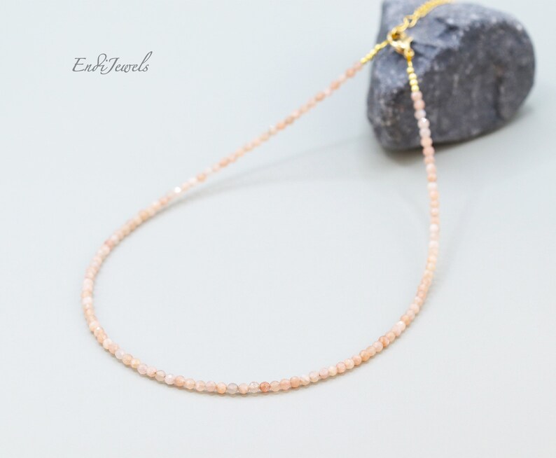 K&ouml;nnte beinhalten: Eine zarte goldene Kettenkette mit kleinen, hellrosa Sunstone-Perlen. Die Kette wird auf einem grauen Stein pr&auml;sentiert.