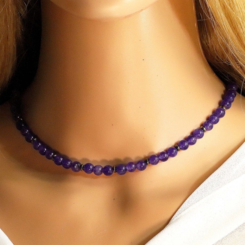 K&ouml;nnte beinhalten: Eine Halskette aus violetten Amethyst-Edelsteinen mit silbernen Perlen. Die Halskette befindet sich auf einer wei&szlig;en Puppe.