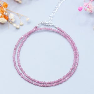May include: A delicate pink gemstone necklace with a silver clasp. The necklace is composed of small, round, pink beads. The background is a light blue with floral accents and the text "Endi Jewels".