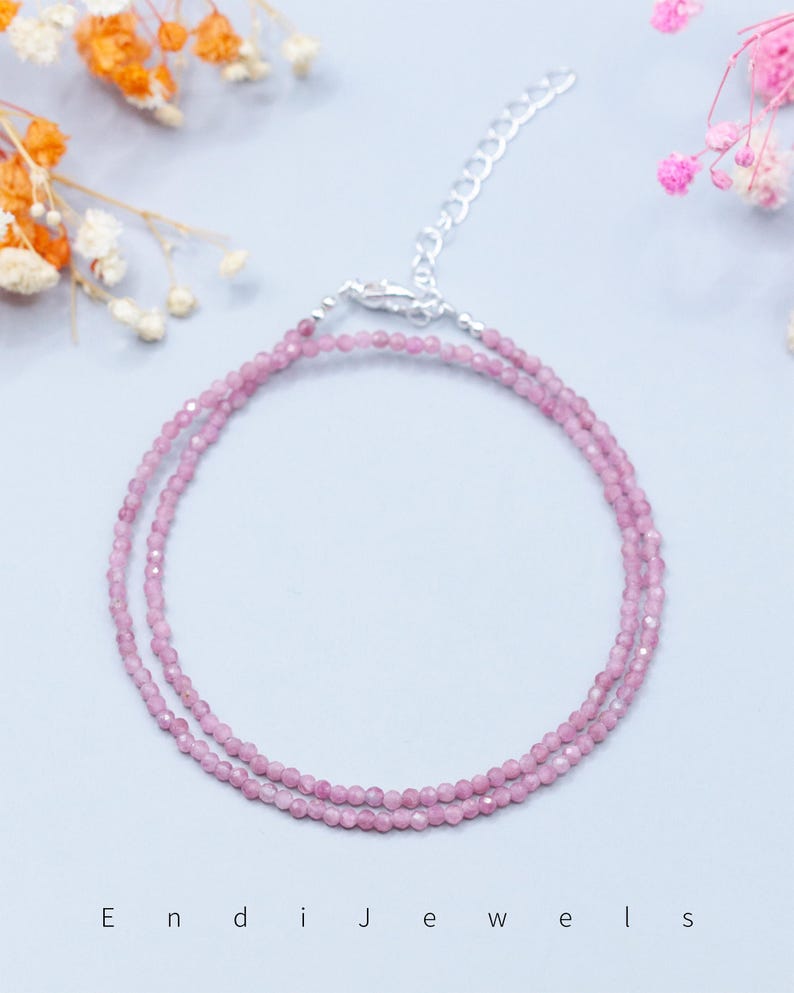 May include: A delicate pink gemstone necklace with a silver clasp. The necklace is composed of small, round, pink beads. The background is a light blue with floral accents and the text "Endi Jewels".
