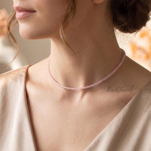 May include: A delicate choker necklace featuring a single strand of small, smooth, light pink beads. The necklace sits closely around the neck and includes the text EndiJewels printed in a cursive script near the collarbone area of the garment.