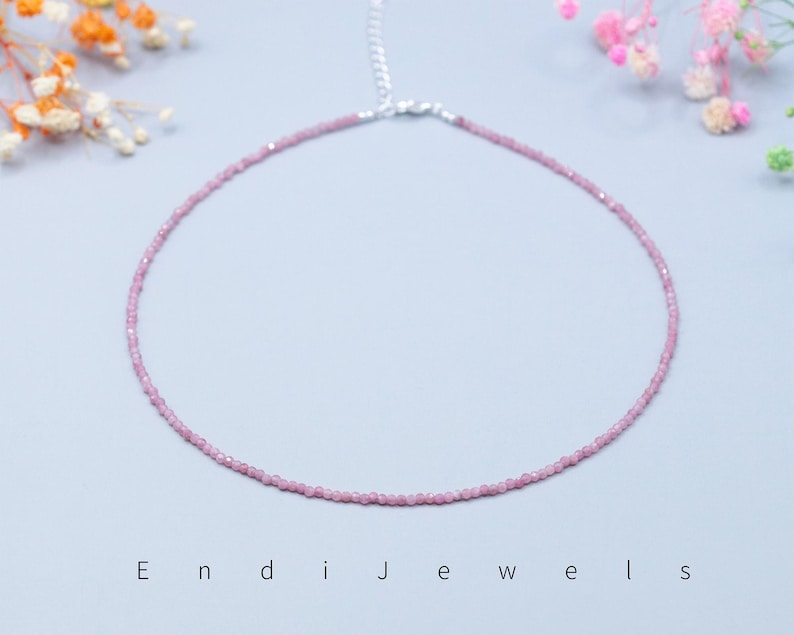 May include: A delicate pink gemstone necklace with a silver clasp. The necklace is composed of small, evenly sized beads, creating a continuous, circular design. The background is a soft, light blue, with floral accents in the corners. The text "Endi Jewels" is at the bottom.