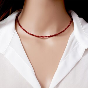 May include: A delicate red beaded necklace. The necklace is composed of small, round beads strung together to form a choker-style design. The beads are a deep red color, and the necklace is worn against a white collared shirt.