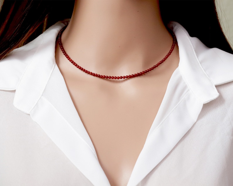 May include: A delicate red beaded necklace. The necklace is composed of small, round beads strung together to form a choker-style design. The beads are a deep red color, and the necklace is worn against a white collared shirt.