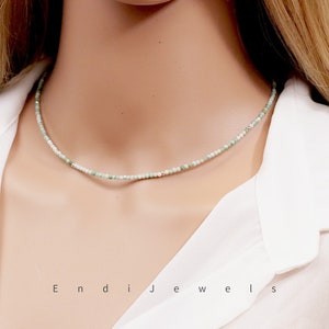 May include: A delicate green and white beaded necklace on a white shirt. The necklace is made of small, round beads and has a simple, elegant design.