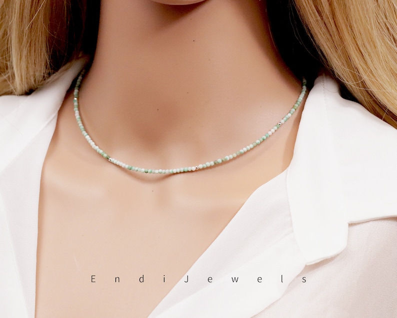 May include: A delicate green and white beaded necklace on a white shirt. The necklace is made of small, round beads and has a simple, elegant design.