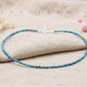 May include: Delicate turquoise beaded necklace with a silver clasp. The necklace is composed of small, round, light blue beads. The word "Endi Jewels" is visible in the background. The necklace is displayed on a neutral-colored fabric.
