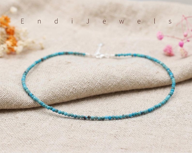 May include: Delicate turquoise beaded necklace with a silver clasp. The necklace is composed of small, round, light blue beads. The word "Endi Jewels" is visible in the background. The necklace is displayed on a neutral-colored fabric.