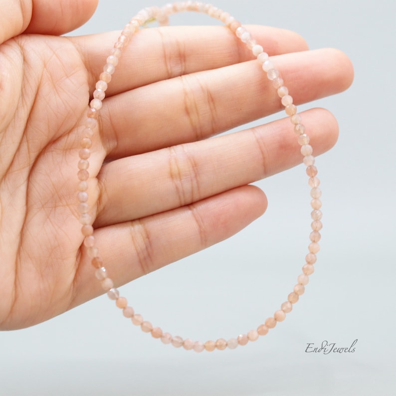 K&ouml;nnte beinhalten: Eine zarte Halskette aus kleinen, hellrosa Sunstone-Perlen. Die Halskette wird auf einer Hand pr&auml;sentiert, wodurch ihre L&auml;nge und der subtile Schimmer der Steine zur Geltung kommen.
