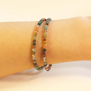 May include: A multi-colored beaded bracelet with green, brown, orange, and red beads. The bracelet is made of small, round beads and is worn on a person's wrist.