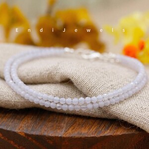 May include: A delicate, light blue beaded necklace with a silver clasp. The necklace is composed of small, round beads and is displayed on a beige linen cloth. The word "Endi Jewels" is visible in the background.