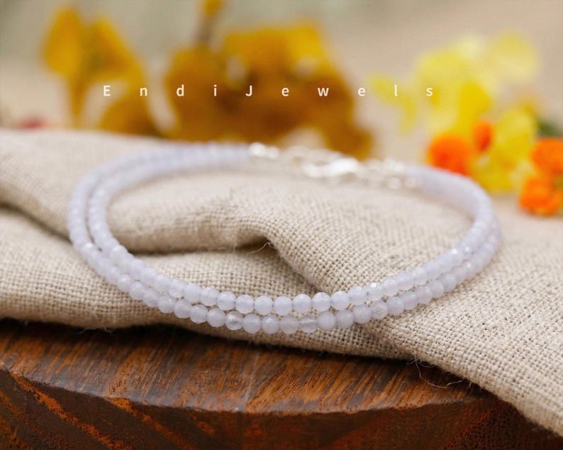 May include: A delicate, light blue beaded necklace with a silver clasp. The necklace is composed of small, round beads and is displayed on a beige linen cloth. The word "Endi Jewels" is visible in the background.