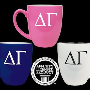 May include: Three ceramic mugs in pink, blue, and white, each with a white or black Greek letter design. The pink mug is at the top, the blue mug is on the left, and the white mug is on the right. The mugs have a curved handle and a glossy finish.