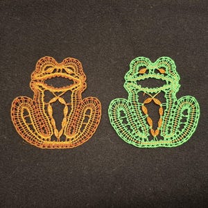 Puede incluir: Dos adornos de rana de ganchillo, uno en naranja y verde, el otro en verde y marrón.