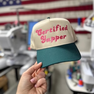 May include: A beige and green baseball cap with the text "Certified Yapper" embroidered in pink.