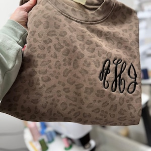 May include: A brown leopard print sweatshirt with black embroidered initials. The sweatshirt features a repeating pattern of leopard spots. The initials are in a cursive font.