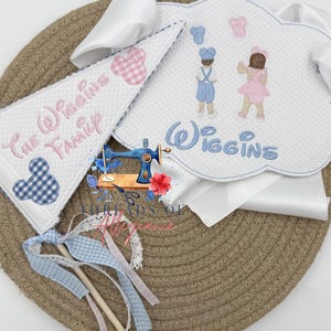 May include: A collection of personalised baby accessories, including a bib, pennant, and wand. The bib features embroidered figures and the name "Wiggins." The pennant displays "The Wiggins Family." The wand has ribbons and lace. Colours include white, blue, pink, and gingham patterns.