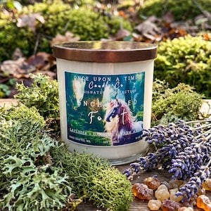 May include: A white soy candle with a copper lid, labeled "Enchanted Forest" with a unicorn illustration. The candle is surrounded by moss, lavender, and amber-colored resin. The candle is 10 oz.