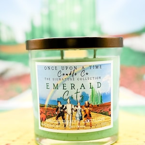 Puede incluir: Un tarro de vela de vidrio verde con tapa dorada. La etiqueta dice "Once Upon A Time Candle Co. The Signature Collection Emerald City" y presenta una escena de El Mago de Oz con un arcoíris y el camino de ladrillos amarillos. El aroma de la vela es "Lima, Pepino y Melón."