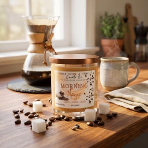 May include: A glass jar candle with a copper lid, labelled "Morning Magic" with coffee bean and sugar cube accents. The candle sits on a wooden surface with coffee beans and sugar cubes scattered around it. A coffee maker and mug are in the background.