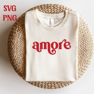 May include: A white t-shirt with the word "amore" in red, retro-style lettering. The shirt is displayed on a woven straw mat.
