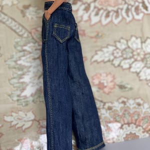 Op de afbeelding: Donkerblauwe denim poppenbroek met gele stiksels. De broek heeft een klassiek vijf-zakken ontwerp en een rechte pijp. De pop draagt ook een top met patroon.