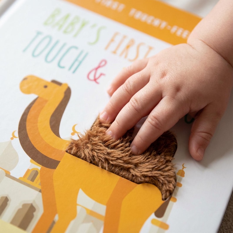 May include: A children's touch-and-feel book with the text "BABY'S FIRST TOUCH &" and a camel illustration. The camel has a brown, textured patch. A child's hand is touching the camel's fur.