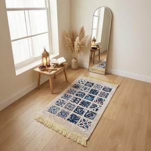 Può includere: Un tappeto da preghiera beige con un motivo a piastrelle geometriche nei toni del blu, rosso e bianco. Il tappeto ha un bordo con frange ed è posizionato su un pavimento in legno. Uno specchio e un tavolino con una lanterna sono sullo sfondo.