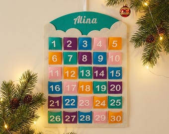 Op de afbeelding: Een vilt adventskalender met gekleurde vakjes genummerd 1-30. De bovenkant is turquoise met de naam "Alina" in witte letters. De kalender is versierd met een kerstboomtak en ornamenten.