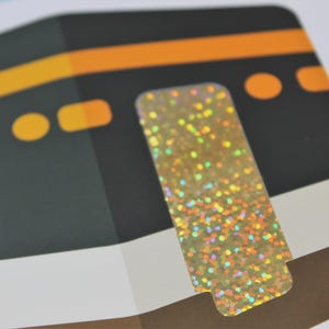May include: Close-up of a card featuring a stylized illustration of the Kaaba, a cube-shaped building in Mecca. The image includes a gold, glittery sticker. The Kaaba is depicted in black with orange and yellow accents.