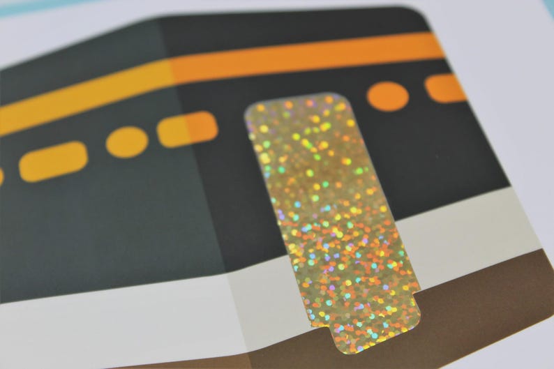 May include: Close-up of a card featuring a stylized illustration of the Kaaba, a cube-shaped building in Mecca. The image includes a gold, glittery sticker. The Kaaba is depicted in black with orange and yellow accents.