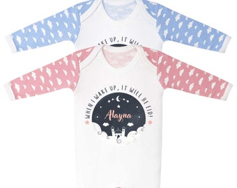 Op de afbeelding: Witte baby romper met roze en blauwe wolken gepatroneerde mouwen. De romper heeft een zwarte cirkel met witte sterren en een halve maan, een moskee silhouet en de tekst "Wanneer ik wakker word, is het Eid!" met de naam "Alayna" in roze.