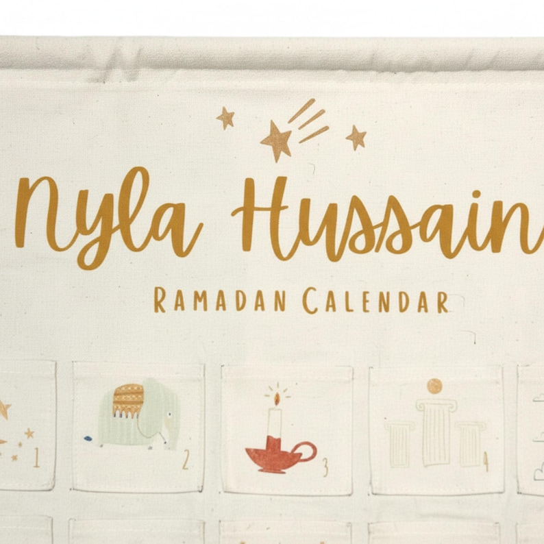 May include: Cream-colored Ramadan calendar with the name "Nyla Hussain" in gold script. The words "Ramadan Calendar" are printed below. The calendar has small pockets with illustrations of stars, an elephant, a candle, and columns.