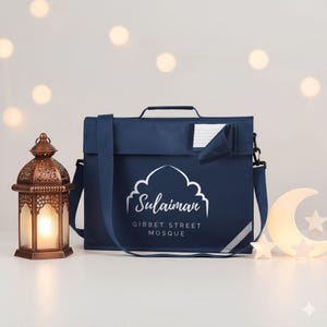 May include: Navy blue school satchel with a white mosque design and the name "Sulaiman" printed on the front. The bag has a shoulder strap, a handle, and a reflective stripe. A decorative lantern and a crescent moon with stars are on the sides.