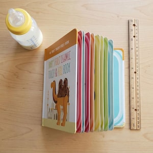 May include: A colorful children's touch-and-feel book titled "Baby's First Islamic Touch & Feel Book" with a camel illustration. A ruler shows the book is approximately 10 inches tall. A baby bottle with milk is also present.