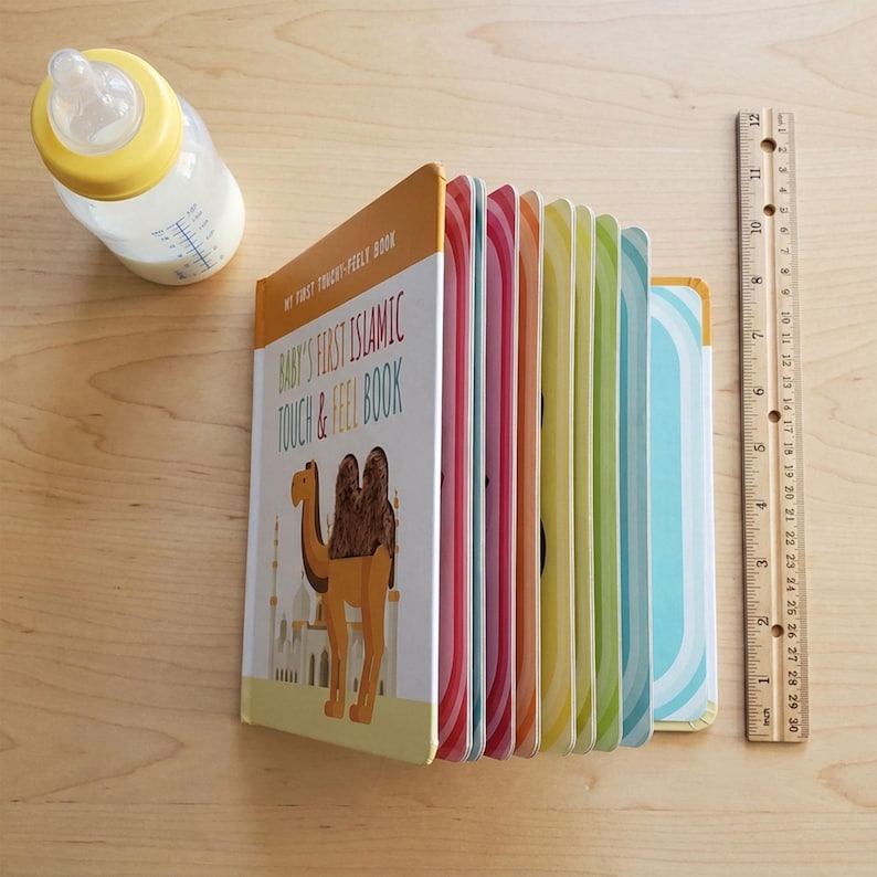 May include: A colorful children's touch-and-feel book titled "Baby's First Islamic Touch & Feel Book" with a camel illustration. A ruler shows the book is approximately 10 inches tall. A baby bottle with milk is also present.