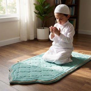 May include: A young child in white traditional clothing kneels on a light teal prayer rug with a quilted design and a tan border. The child is wearing a white cap and has their hands raised in prayer.
