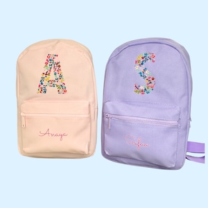 Può includere: Due zaini personalizzati, uno rosa con la lettera A e il nome Anaya, e uno viola con la lettera S e il nome Safaa. Entrambi gli zaini hanno ricami floreali.