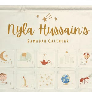 May include: Ramadan calendar with the text "Nyla Hussain's Ramadan Calendar" in gold script. The calendar has small pockets with illustrations of a crescent moon, elephant, candle, and other festive images.