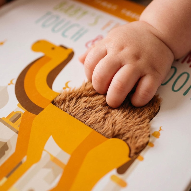 May include: A baby's hand touches a camel illustration in a children's book. The camel is depicted in shades of orange and brown, with a textured, furry patch representing its hump. The book's title includes the words "Baby's First Touch & Feel".