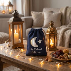 May include: A navy blue drawstring gift bag with a white crescent moon and star design, and the words "Eid Mubarak Eshaan". The bag sits on a wooden table with lit lanterns, fairy lights, and a plate of dates and nuts.