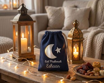 Op de afbeelding: Een marineblauwe geschenkzak met trekkoord, versierd met een witte maan en ster, en de tekst "Eid Mubarak Eshaan". De zak staat op een houten tafel met verlichte lantaarns, lichtjes en een bord met dadels en noten.