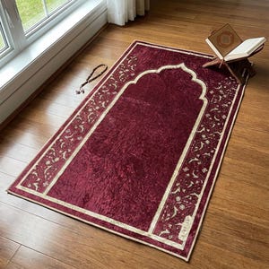 May include: A burgundy prayer rug with a decorative border featuring floral patterns. The rug has a central arch design and is placed on a wooden floor. A wooden book stand with an open book and a string of prayer beads are also present.